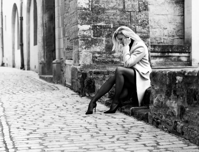 Séance photo urbaine noir et blanc, femme blonde en manteau clair assise sur des marches en pierre